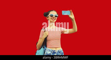 Porträt einer glücklichen lächelnden jungen Frau, die Selfie mit Smartphone macht und coole Girly Hairstyle trägt Rucksack auf rotem Hintergrund Stockfoto