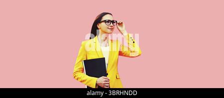 Porträt der eleganten jungen Frau mit Ordner, der wegschaut, Brille trägt, gelber Business-Anzug auf rosa Studiohintergrund Stockfoto