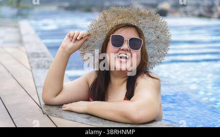 Übergewichtige junge asiatische Frau trägt orange Badeanzüge und Strohhut am Strand, der sich im Pool entspannt, glückliche und große Frau, fröhlich lustiger Urlaub, der im Hotel reist Stockfoto