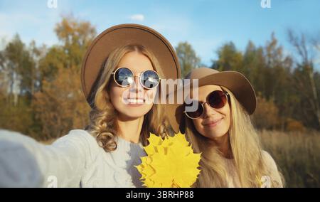 Porträt von glücklichen jungen Freundinnen, die Selfie mit Smartphone machen, das herbstgelbe Blätter hält, stilvolle, lächelnde Freundinnen im Freien, ich Stockfoto