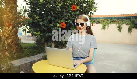 Glückliche junge Frau, die mit einem Laptop arbeitet und Musik in Kopfhörern hört, die am Tisch im Straßencafé im Sommerpark sitzt Stockfoto