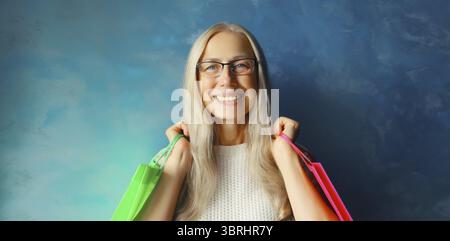 Porträt der schönen glücklichen lächelnden Reifen Frau mit bunten Einkaufstaschen in der Brille auf blauem Hintergrund Stockfoto