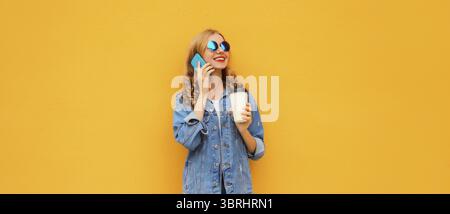 Moderne glückliche junge Frau, die auf Handy in Denim-Jacke auf gelbem Studiohintergrund anruft Stockfoto