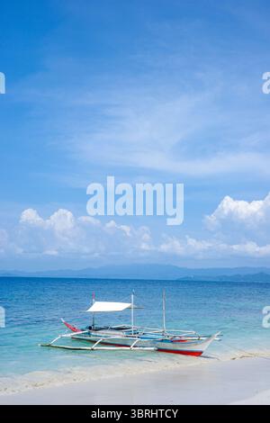Ein traditionelles philippinisches Auslegerboot oder bangka liegt an einem ruhigen weißen Sandstrand unter einem blauen Himmel mit türkisfarbenem Wasser über der Küste Stockfoto