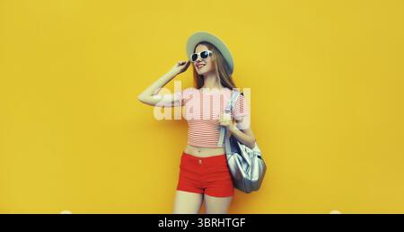Schöne junge Frau, die im Sommer Strohhut posiert, rote Shorts mit Rucksack, die auf lebhaftem gelbem Studiohintergrund wegschauen Stockfoto