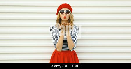 Porträt der schönen glücklichen jungen Frau, die einen Kuss mit Lippenstift in roter französischer Baskenmütze bläst, herzförmige Sonnenbrille auf weißem Hintergrund Stockfoto