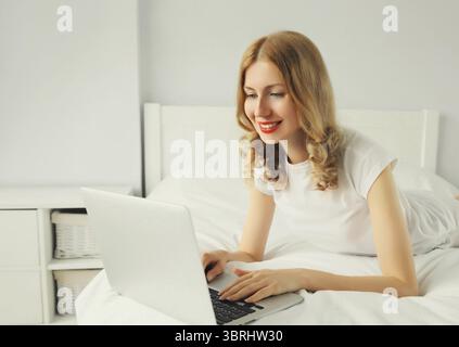 Porträt einer lächelnden jungen Frau, die mit einem Laptop auf dem Bett im weißen Zimmer zu Hause arbeitet Stockfoto