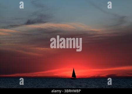Ein einsames Segelboot treibt am TLV Beach über das Mittelmeer, während ein feuriger Sonnenuntergang den Horizont übermalt und eine ruhige Silhouette über sanften Wellen wirft. Stockfoto