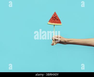 Sommerbild einer Frau mit süßem Lutscher oder eisförmigem Stück Wassermelone, weibliche Hände zeigen saftige Früchte auf blauem Hintergrund Stockfoto