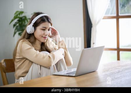 Glückliche schöne asiatische Frau, die Kopfhörer mit Laptop bei der Arbeit trägt oder Musik genießt Stockfoto