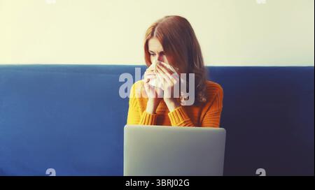 Porträt einer traurigen, kranken, verärgerten Frau, die niesend Nasenschnase mit Taschentüchern beim Arbeiten am Laptop auf dem Sofa zu Hause sitzt Stockfoto