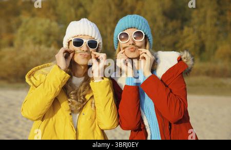 Porträt von glücklichen zwei Freundinnen, die Spaß haben, Schnurrbart in winterlichem Hut, Brille und lustigen Freundinnen im Park zu zeigen Stockfoto
