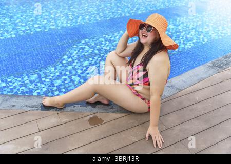 Übergewichtige junge asiatische Frau trägt orange Badeanzüge und Strohhut am Strand, der sich im Pool entspannt, glückliche und große Frau, fröhlich lustiger Urlaub, der im Hotel reist Stockfoto