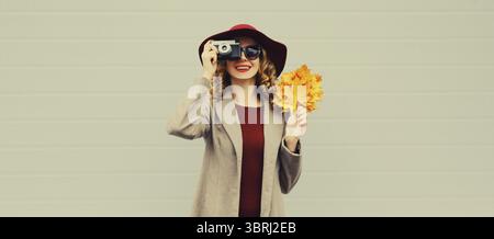 Herbstfarben-Outfit, schöne elegante Fotografin, die mit der Kamera fotografiert, hält gelbe Ahornblätter mit Hut auf grauem Hintergrund Stockfoto