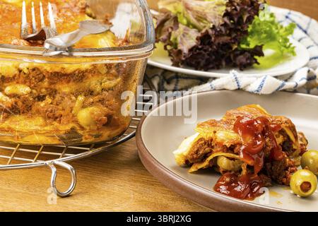 Ein Stück köstliche hausgemachte Lasagne in brauner Keramikschale mit Lasagne in transparenter Glasschale auf einem Kühlregal und frisches Gemüse auf einer weißen Platte Stockfoto