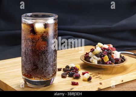 Nahaufnahme von kaltem Cola-Wasser mit Eiswürfeln in transparentem Glas und verschiedenen getrockneten tropischen Früchten in Holzschale auf Holzschneidebrett auf schwarzem Backgr Stockfoto