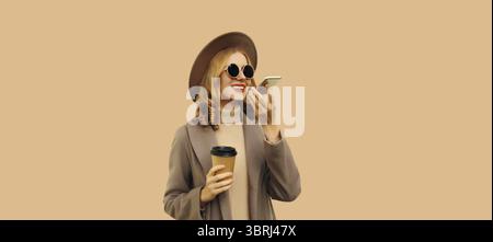 Schöne lächelnde junge Frau mit Kaffeetasse, die Handy mit Sprachaufzeichnungsgerät hält, Assistent oder nimmt Anrufe mit braunem Mantel, rundem Hut Stockfoto