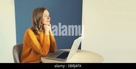 Moderne junge Frau, die mit einem Laptop arbeitet, sitzt am Schreibtisch im Zimmer zu Hause und blickt nachdenklich aus dem Fenster Stockfoto