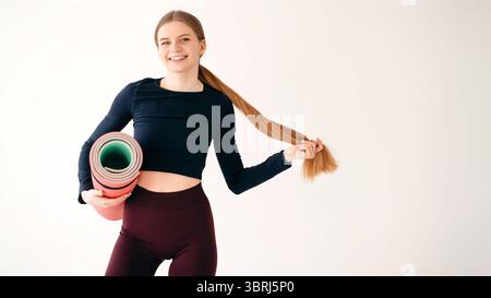 Porträt einer fröhlichen Fitness-Dame, die auf der Kamera mit Gummimatte posiert und glücklich lächelnd auf weißem Hintergrund. Emotionale Frau, die den Pferdeschwanz in der Hand hält Stockfoto