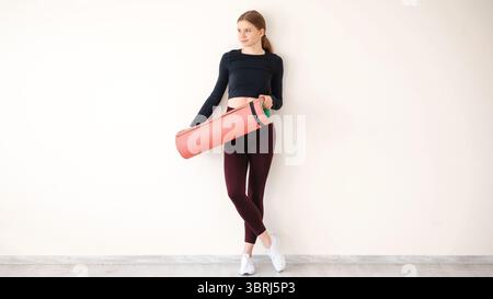 Ganzkörperaufnahme einer schlanken Frau mit Fitness-Körper, trägt Oberteil, Leggings und Sneakers, hält Trainingsmatte, wird im Fitnessstudio trainieren, stellt sich gegen gr Stockfoto