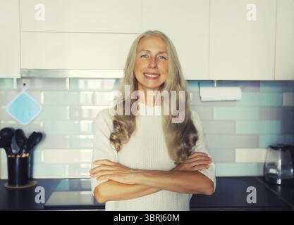 Porträt einer glücklichen lächelnden selbstbewussten Hausfrau mittleren Alters mit grauhaarigen Armen in der Küche zu Hause Stockfoto
