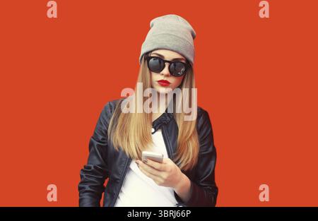 Porträt einer stilvollen Frau mit Smartphone mit grauem Hut, Leder-Biker-Rock-Jacke auf rotem Hintergrund Stockfoto