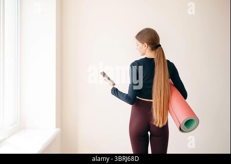 Frau mit Pferdeschwanz, die beim Training oder Yoga-Kurs stand, mit einer rollenden Fitnessmatte und einem Smartphone in ihrem Mann drehte sie der Kamera den Rücken zu. Speicherplatz kopieren auf Stockfoto