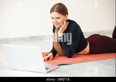 Lächelnde langhaarige Frau, die auf dem Bauch auf roter Yogamatte liegt und auf den Laptop-Bildschirm im weißen Aerobic-Studio blickt. FIT-Dame auf der Suche nach motivierender tr Stockfoto