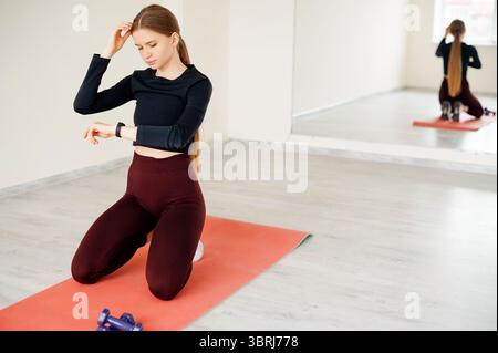 Winkelansicht einer erwachsenen Frau, die auf Knien auf roter Gummimatte sitzt und mit Kurzhanteln auf Smartwatch-Display blickt. Gesunde Frau, die den Puls bei Fitnes überprüft Stockfoto