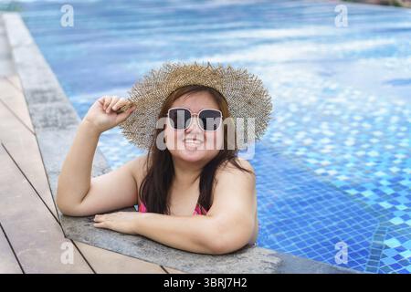 Übergewichtige junge asiatische Frau trägt orange Badeanzüge und Strohhut am Strand, der sich im Pool entspannt, glückliche und große Frau, fröhlich lustiger Urlaub, der im Hotel reist Stockfoto
