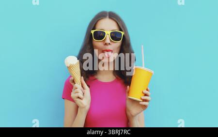 Sommerporträt einer glücklichen jungen Frau, die Eis isst und eine Tasse frischen Saft hält, die Sonnenbrille auf blauem Hintergrund trägt Stockfoto