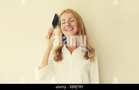 Porträt einer glücklichen lächelnden kaukasischen blonden jungen Frau, die ihr Haar mit Kamm auf weißem Studiohintergrund kämmt Stockfoto