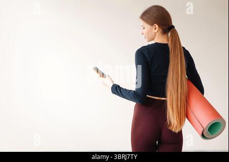 Frau mit Pferdeschwanz, die beim Training oder Yoga-Kurs stand, mit einer rollenden Fitnessmatte und einem Smartphone in ihrem Mann drehte sie der Kamera den Rücken zu. Speicherplatz kopieren auf Stockfoto