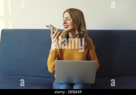 Moderne junge Frau, die mit einem Laptop arbeitet, telefoniert oder mit einem Sprachassistenten, Kommandorecorder sitzt auf einer Couch im Wohnzimmer bei Stockfoto