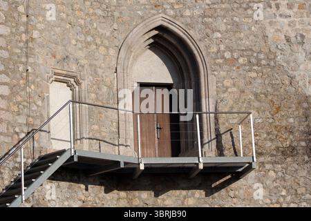 Ein spitzer gotischer Bogengang mit Holztüren öffnet sich auf einen modernen Metallbalkon, der den Kontrast zwischen mittelalterlicher Handwerkskunst unterstreicht Stockfoto