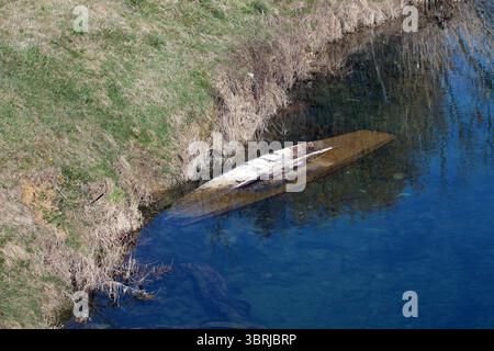 Ein altes Holzboot liegt umgekippt und teilweise in transparentem, seichten Wasser in der Nähe eines grasbewachsenen Ufers, dessen Rumpf sichtbar verwittert ist Stockfoto
