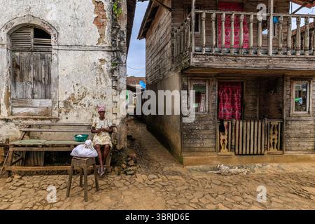 Ein einheimischer Dorfbewohner sitzt auf einer Holzbank vor einem heruntergekommenen Kolonialgebäude und neben einem traditionellen Holzhaus in einer Kopfsteinpflasterstraße in sao Tome Stockfoto