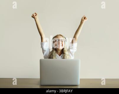 Glückliche, freudige Frau mittleren Alters, die Musik in Kopfhörern hört und mit einem Laptop arbeitet, der zu Hause am Schreibtisch sitzt Stockfoto
