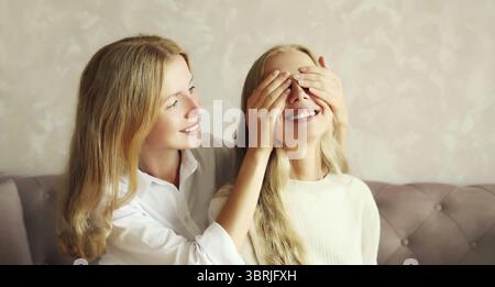 Glückliche lächelnde weiße erwachsene Tochter, die die Augen der Mutter bedeckt, überraschend auf der Couch zu Hause sitzend Stockfoto