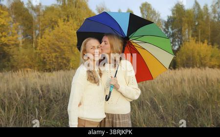 Erwachsene Tochter küsst ihre glückliche, lächelnde, reife Mutter mit buntem Regenschirm, der mit gelben Blättern im Herbstpark spaziert Stockfoto