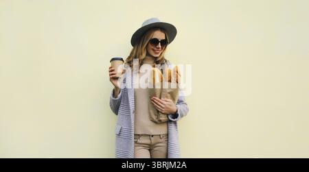Porträt der schönen glücklichen jungen Frau, die Lebensmitteleinkaufpapiertüte mit weißem Brot Baguette hält, der Kaffee nach der Bäckerei auf weißem Hintergrund trinkt Stockfoto