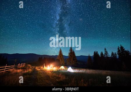 Verschwommene Silhouetten von Touristen, die auf dem Campingplatz am Lagerfeuer ruhen, mit leuchtenden Zelten in der Nähe unter Sternenhimmel. Die Milchstraße erstreckt sich über die Berge und wirft magisches Licht über die beschauliche Berglandschaft. Stockfoto