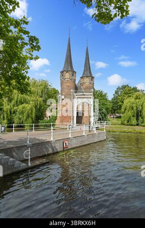 Das historische Oostpoort (östliches Tor) in Delft, Niederlande, mit seinen Doppeltürmen, der Zugbrücke und dem ruhigen Kanal, eingerahmt von üppigen Bäumen. Stockfoto