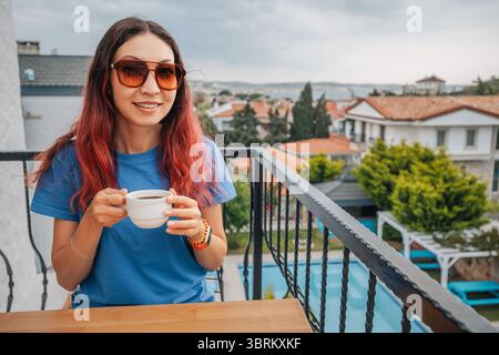 Junge Frau mit Sonnenbrille genießt Morgenkaffee auf dem Balkon mit Blick auf die mediterrane Stadt mit Swimmingpool Stockfoto