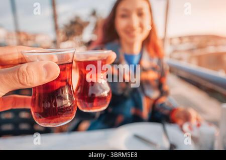 Zwei türkische Teegläser stoßen bei einem Toast an, während ein lächelnder Tourist den Moment vor einer lebhaften Kulisse bei Sonnenuntergang genießt Stockfoto