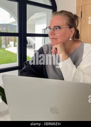 Faro, Portugal - 13. Juli 2025: Frau, die in einem eleganten Home Office mit hellen Fenstern und üppigem Garten unterwegs ist, arbeitete an einem Apple-Laptop Stockfoto