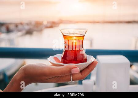 Frau Hand hält eine Tasse traditionellen türkischen Tee mit einer verschwommenen Meereslandschaft im Hintergrund bei Sonnenuntergang Stockfoto