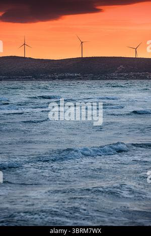 Windturbinen drehen sich auf einem Hügel und erzeugen saubere Energie, während ein atemberaubender Sonnenuntergang den Himmel in leuchtenden Orange-Tönen über dem Meer überzieht Stockfoto