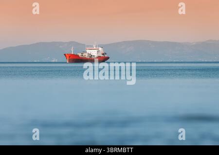 Frachtschiff segelt auf ruhigem Meer bei Sonnenuntergang mit Bergen im Hintergrund, Seeverkehrskonzept Stockfoto