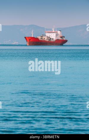 Rotes Frachtschiff auf ruhigem blauem Meer mit Bergen im Hintergrund, Seeverkehrskonzept Stockfoto